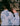 A woman stands with her back to the camera. She is wearing Saywood's Marie A-line Blouse in pale blue recycled cotton, trimmed with a narrow lace trim and gathered frill cuffs. She is plaiting her hair, whilst shadows of the trees are visible on her back.