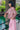 A woman of Japanese heritage is leaning with one arm on an iron railing, overlooking the Barbican pond, with the building just visible in the background. She is wearing Saywood's pink and hazelnut check Marie A-line Blouse with gathered neck details and gathered frill cuffs, with the Amelia Wide Leg Culotte Trousers in pink cotton corduroy. She has one her hand in her pocket, and wears white sunglasses with pink lenses.