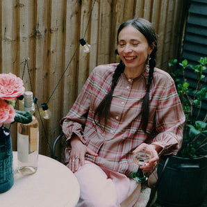 A woman is sitting in a iron garden chair holding a glass of wine, with a small round marble table in front of her, with a bottle of wine on it and a blue vase with large pink rose flowers. She is wearing Saywood's pink and hazelnut check Marie A-line Blouse with gathered neck details and gathered frill cuffs, with the Amelia Wide Leg Culotte Trousers in pink cotton corduroy. She's smiling, with two plaited pigtails in her hair. Behind her is a wooden fence with outdoor lights strung to it and a plant.