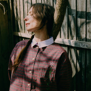 Close up of a woman leaning against a tree in front of a wooden fence. She has her eyes closed and is dappled in sunlight. She wears Saywood's red check Etta Oversized Shirtdress with signature embroidered flower on the pocket, and lilac contrast collar.
