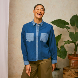 Model stands in front of a pink drop of fabric and two plants in the background. She wears Saywood's Japanese Denim Lela Patchwork Shirt, with light wash contrast cuffs, pockets and collar. Worn with khaki Amelia Trousers.