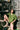 A woman is perched on a garden pot, holding a garden hose, with a wooden fence behind her, with leafy shadows on the fence. She is wearing the Frances Unisex Cuban Shirt in olive green, with short sleeves and the signature embroidered flower in lilac on the check pocket. Worn with the Bessie A-line Shorts in olive green as a matching twin set. She is holding a curled up garden hose, with one leg up on the garden chimney pot.