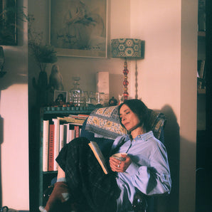 A woman sits in a blue antique armchair, with her knees up, holding a cup of tea with a book in her lap. She is wearing Saywood's Edi Volume Sleeve Shirt with lace trim collar in pale blue recycled cotton, with the Amelia Wide Leg Trousers in navy check, and cosy brown and blue socks. A bookcase is behind her, with a lamp and ornaments on, with a picture by artist Linder Sterling on the wall.