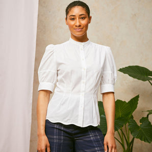 Model stands smiling, wearing Saywood's Joni puff sleeve blouse in white with scalloped hem, worn with navy check Amelia wide leg trousers. A plant and drop of pink fabric can be seen in the background.