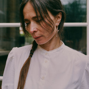 Close up of a woman with dark hair, plaited, looking to one side. She is wearing Saywood's Joni Puff Sleeve Blouse, with a silver twisted earring in her ear. The buttons down the blouse can be seen, as well as the grandad collar with lace trim on the edging, and puff sleeve detailing.