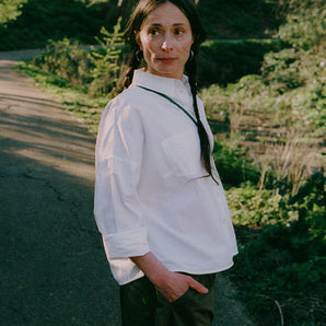 A woman is standing on a path in a green park surrounded by nature. She is turned to the side looking over her shoulder with her hand in her trouser pocket. She wears Saywood's white recycled cotton Lela Boxy Shirt with the khaki Amelia Wide Leg Trousers. Her hair is plaited in pigtails and she has a green cross body bag across her.