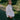 A woman is standing on a path in a green park surrounded by nature. She is turned to the side looking over her shoulder with her hand in her trouser pocket. She wears Saywood's white recycled cotton Lela Boxy Shirt with the khaki Amelia Wide Leg Trousers. Her hair is plaited in pigtails and she has a green cross body bag across her.