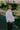 A woman is standing on a path in a green park surrounded by nature. She is turned to the side looking over her shoulder with her hand in her trouser pocket. She wears Saywood's white recycled cotton Lela Boxy Shirt with the khaki Amelia Wide Leg Trousers. Her hair is plaited in pigtails and she has a green cross body bag across her.