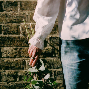 Close up of the arm and body of a woman standing in front of a brick wall, holding a bunch of eucalyptuses. She is wearing rings on her fingers, and jeans, worn with Saywood's white recycled cotton Marie A-line Gathered Neck blouse. the sleeve detail of the gathered cuff and frill with lace trim can be seen. Dappled light and leafy shadows are seen on the blouse.