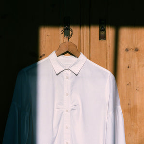 Saywood's Edi Volume Sleeve shirt with lace trim collar is hanging on a wooden hanger on an old French pine wardrobe. The room bathed in sunlight a square of direct sunlight from the window opposite can be seen shining on the shirt, with the surround in darker shade. The soft gathers at the bust on the shirt can be seen as a detail.