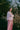 A woman standing amongst the Spring brambles, image shot on film. She wears Saywood's Amelia Wide Leg Trousers in pink corduroy, paired with the Lela Boxy Colourblock Shirt in pastel pink and orange, all made in London. Her hair is in pigtail plaits and her arms are folded across her chest.