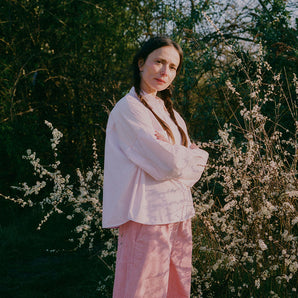 A woman standing amongst the Spring brambles, image shot on film. She wears Saywood's Amelia Wide Leg Trousers in pink corduroy, paired with the Lela Boxy Colourblock Shirt in pastel pink and orange, all made in London. Her hair is in pigtail plaits and her arms are folded across her chest.